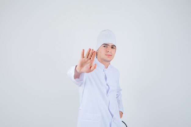 muchacho adolescente mostrando gesto de parada en uniforme de médico y mirando serio