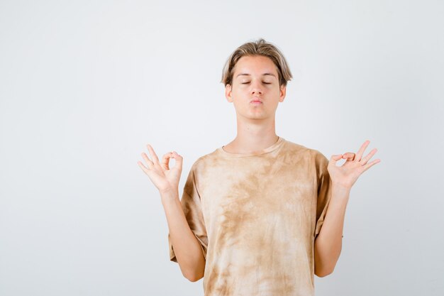 Muchacho adolescente mostrando gestos de yoga, haciendo pucheros con los labios en camiseta y mirando pacífica, vista frontal.