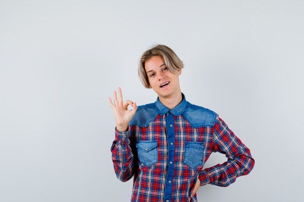 Muchacho adolescente mostrando signo ok en camisa a cuadros y mirando alegre, vista frontal.