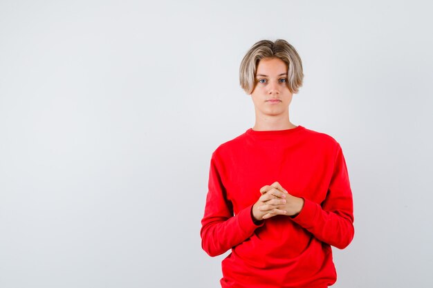 Muchacho adolescente en suéter rojo manteniendo los dedos entrelazados y mirando confiado, vista frontal.