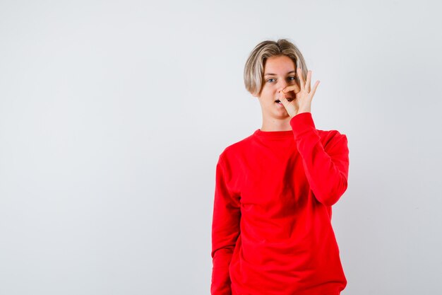 Muchacho adolescente en suéter rojo que muestra un gesto aceptable y que parece alegre, vista frontal.