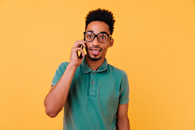 Muchacho asombrado en vasos grandes hablando por teléfono. Retrato interior de chico africano emocional en camiseta verde llamando a alguien.