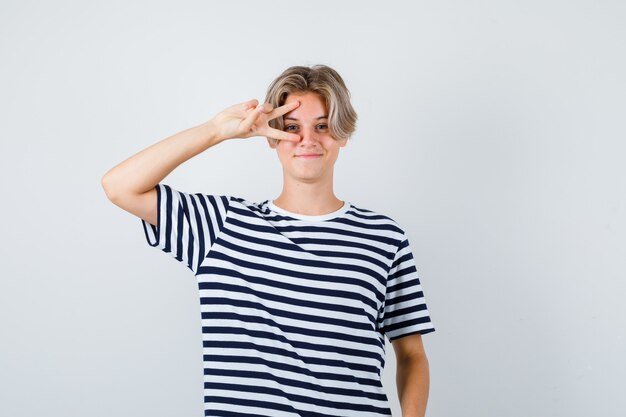 Muchacho bastante adolescente en camiseta rayada que muestra el signo de V cerca del ojo y mirando alegre, vista frontal.
