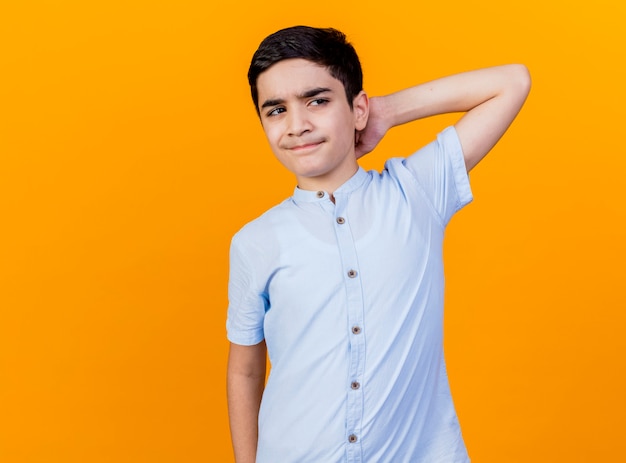 Muchacho caucásico joven confundido que mira al lado manteniendo la mano detrás del cuello aislado sobre fondo naranja con espacio de copia