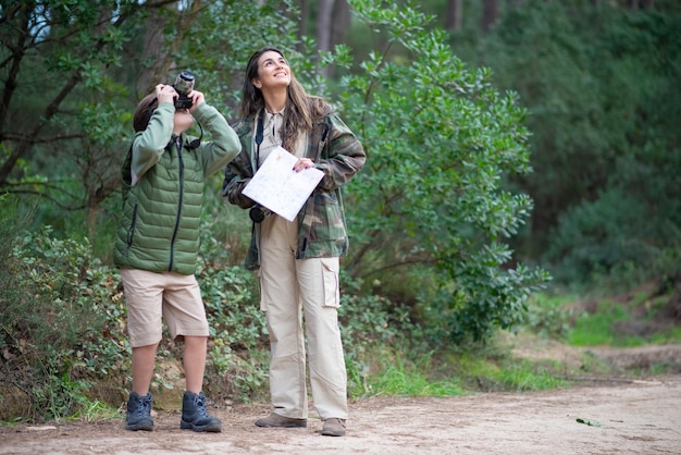 Muchacho feliz y wo tomando fotos en el bosque. Madre e hijo de cabello oscuro, niño apuntando la cámara a los árboles. Crianza, familia, ocio, concepto de hobby.