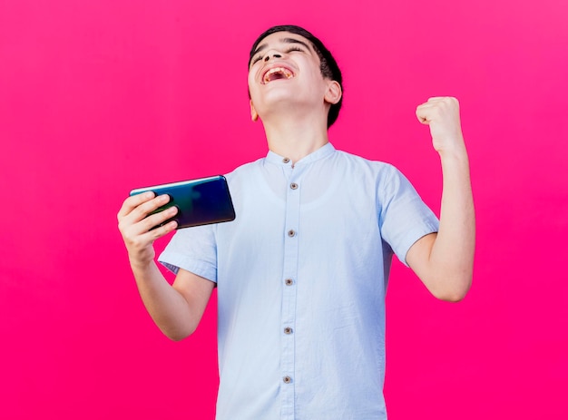 Muchacho joven alegre que sostiene el teléfono móvil que hace sí el gesto con los ojos cerrados aislado en la pared rosada