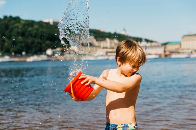 El muchacho joven derrama el agua del cubo en la playa del mar