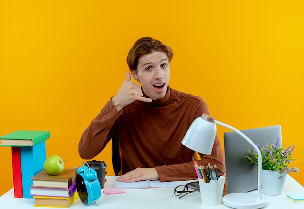 Muchacho joven estudiante alegre sentado en el escritorio con herramientas escolares mostrando gesto de llamada telefónica