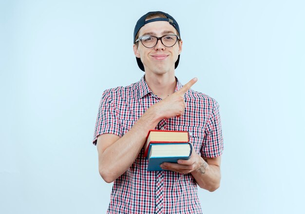 Muchacho joven estudiante complacido con bolsa trasera y gafas y gorra sosteniendo libros y puntos al lado en blanco