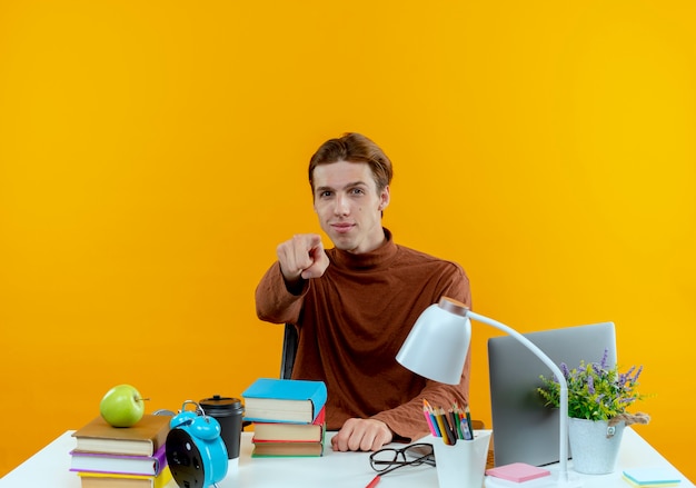 Muchacho joven estudiante complacido sentado en el escritorio con herramientas escolares que le muestran gesto aislado en la pared amarilla