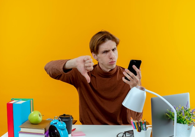 Muchacho joven estudiante descontento sentado en el escritorio con herramientas escolares sosteniendo el teléfono con el pulgar hacia abajo en amarillo