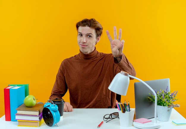 Muchacho joven estudiante satisfecho sentado en el escritorio con herramientas escolares mostrando tres aislados en la pared amarilla