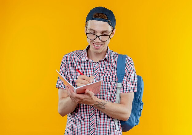 Muchacho joven estudiante sonriente que lleva el bolso trasero y los anteojos y la tapa que escriben algo en el cuaderno en blanco