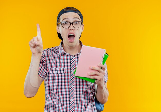 Muchacho joven estudiante sorprendido que lleva el bolso trasero y los anteojos y la tapa que sostiene el cuaderno y apunta hacia arriba en blanco