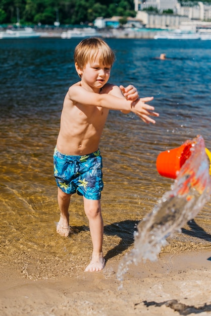 Muchacho joven en pantalones cortos que lanza el cubo con agua en la playa del mar