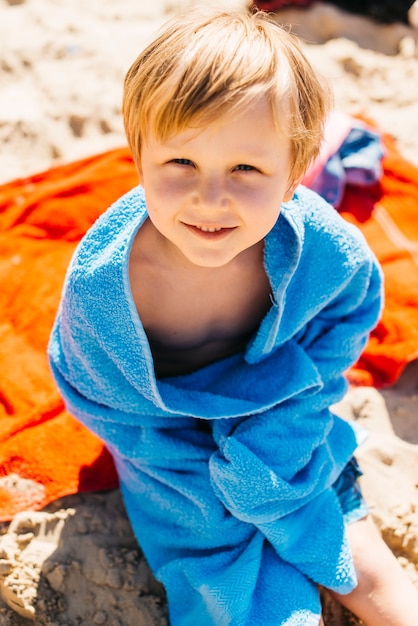 Muchacho joven que se sienta en la playa en toalla azul