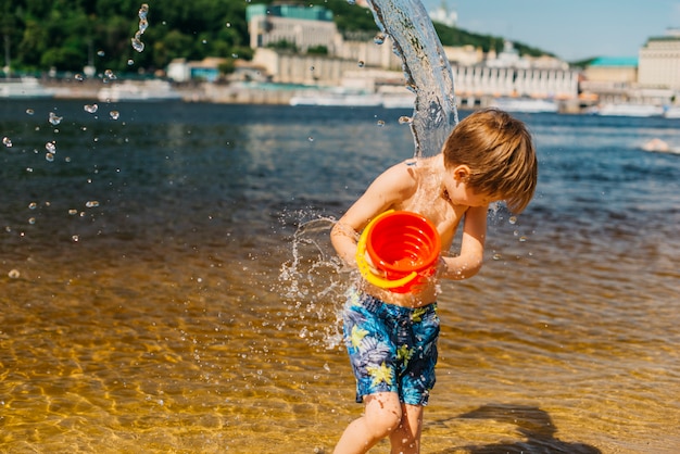 Foto gratuita muchacho joven que se vierte del cubo con agua en la playa del mar