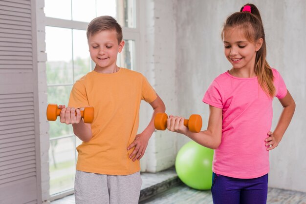 Muchacho y muchacha sonrientes con la mano en las caderas que ejercitan con pesas de gimnasia