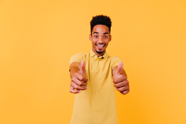 Muchacho negro refinado que muestra los pulgares para arriba con sonrisa sorprendida. Foto interior de hombre africano de buen humor con peinado divertido.