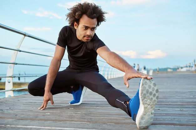 Muchacho de piel oscura atleta sano que se extiende en la plataforma de madera en la mañana. hombre deportivo con peinado tupido calentando sus piernas