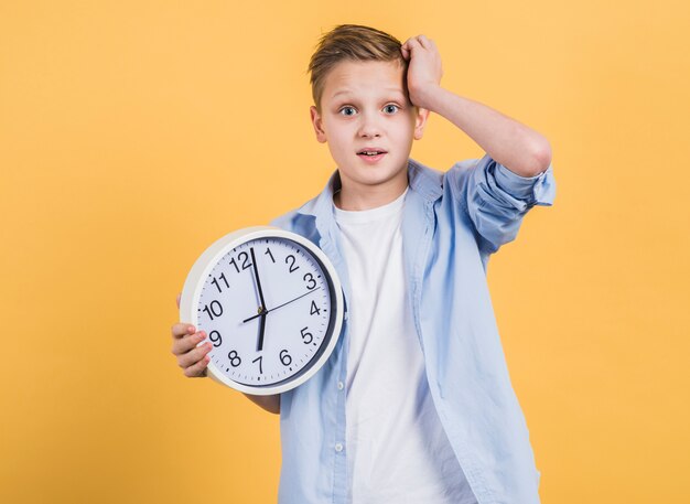 Muchacho preocupado con la mano en su cabeza que sostiene el reloj blanco que se opone a fondo amarillo