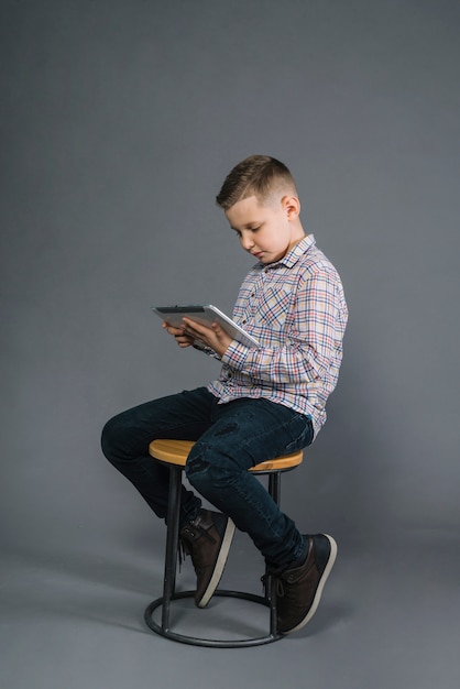 Un muchacho que se sienta en el taburete que mira la tableta digital contra fondo gris