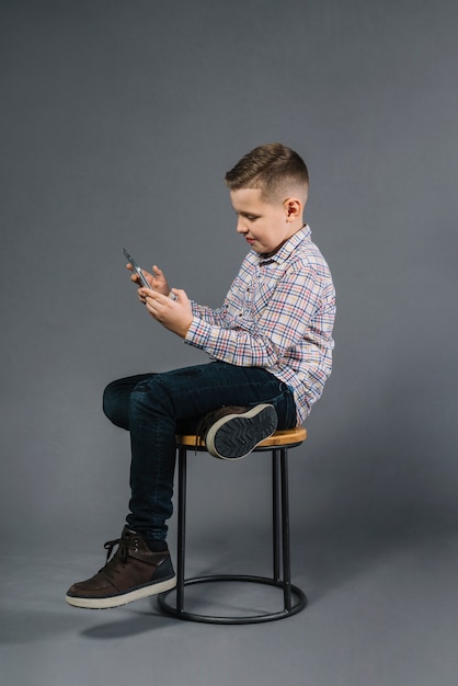 Un muchacho que se sienta en taburete usando el teléfono móvil contra fondo gris