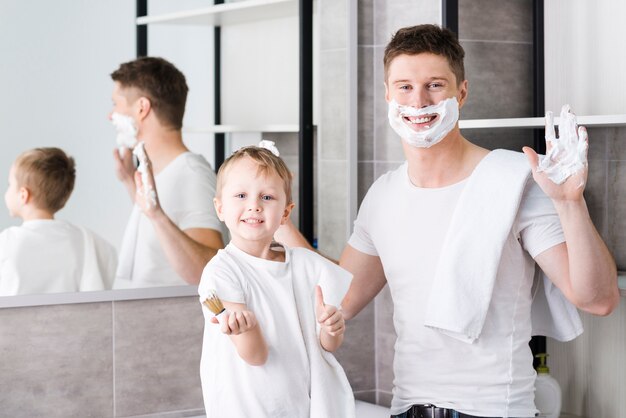 Muchacho que sostiene el cepillo en la mano que muestra el pulgar hacia arriba signo de pie cerca de su padre con espuma de afeitar en su cara y mano