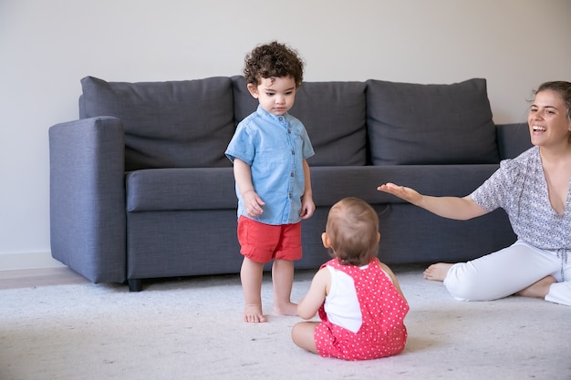 Muchacho de raza mixta serio de pie y mirando al bebé. Mamá bonita caucásica hablando algo con los niños, sonriendo y jugando con los niños en casa. Concepto de familia en el interior, fin de semana e infancia