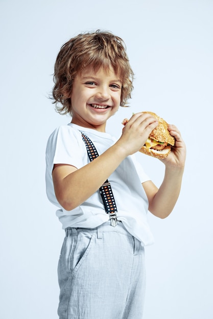 Muchacho rizado bastante joven en ropa casual en la pared blanca. Comer hamburguesa. Niño en edad preescolar masculino caucásico con brillantes emociones faciales. Infancia, expresión, diversión, comida rápida. Sonriente.