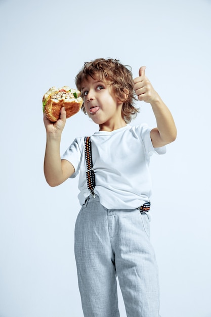 Muchacho rizado bastante joven en ropa casual en la pared blanca. Comer hamburguesa. Niño en edad preescolar varón caucásico con brillantes emociones faciales. Infancia, expresión, diversión, comida rápida. Mostrando el pulgar hacia arriba.