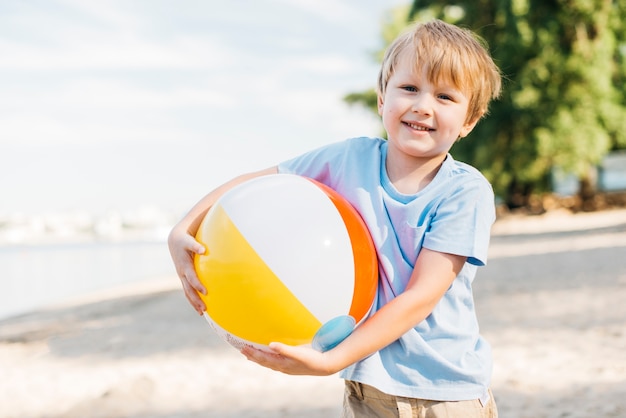 Muchacho sonriente que lleva la pelota de playa ambas manos