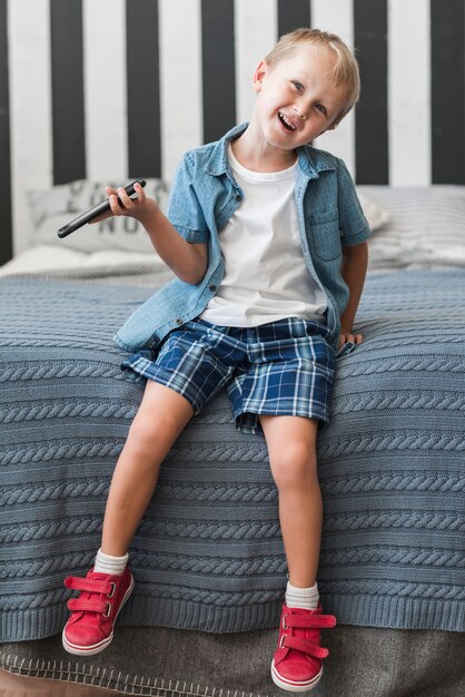 Muchacho sonriente que se sienta en la cama que sostiene el teléfono elegante