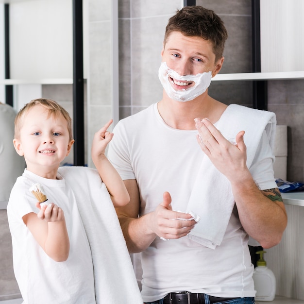 Foto gratuita muchacho sonriente que sostiene la brocha de afeitar en la mano que señala el dedo hacia su padre con espuma que afeita en su cara