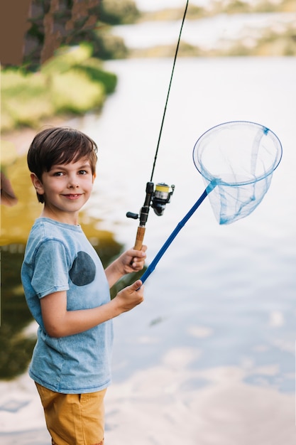 Muchacho sonriente que sostiene la caña de pescar y la red a disposición cerca del lago