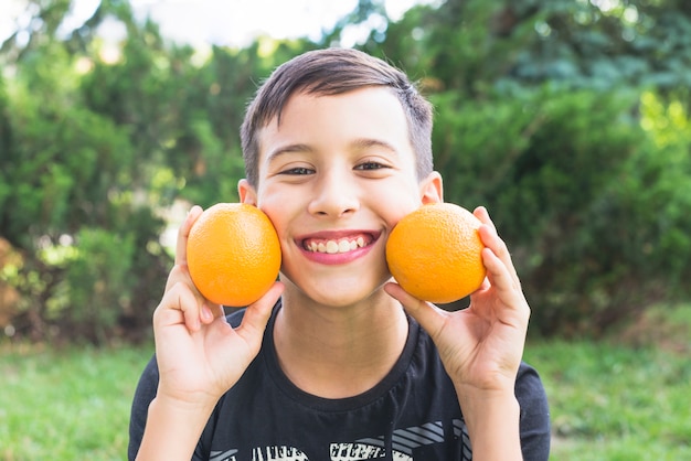 Foto gratuita muchacho sonriente que sostiene naranjas enteras frescas cerca de sus mejillas