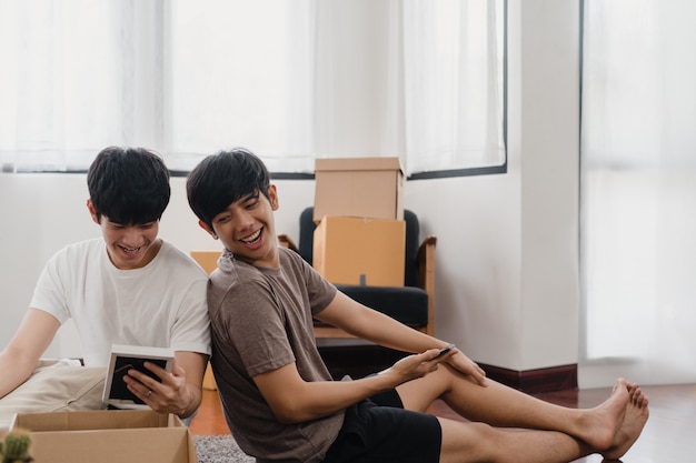 Las mudanzas felices de la reubicación de la pareja gay asiática joven se instalan en un nuevo hogar. Asia lover guy lgbtq + caja de cartón abierta o paquete desempacando en la sala de estar el día de la mudanza. Vivienda inmobiliaria, préstamo e hipoteca.