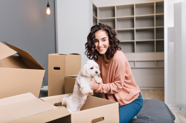 Mudarse al nuevo y moderno apartamento de una joven alegre que encuentra un perrito blanco en una caja de cartón. Sonriendo de hermosa modelo con pelo corto y rizado morena en comodidad de casa