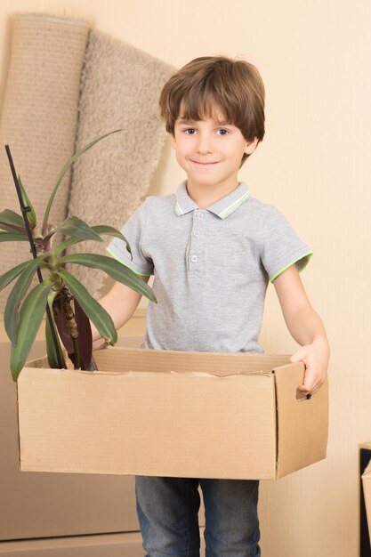 Mudarse a una nueva casa. Retrato de primer plano de niño sonriente feliz sosteniendo una caja con una planta y mirando a la cámara.