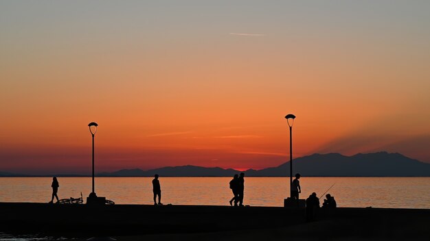 Muelle con varios pescadores y poca gente al atardecer, bicicleta estacionada, farolas de tierra, Grecia