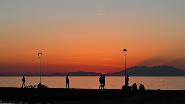 Muelle con varios pescadores y poca gente al atardecer, bicicleta estacionada, farolas de tierra, Grecia