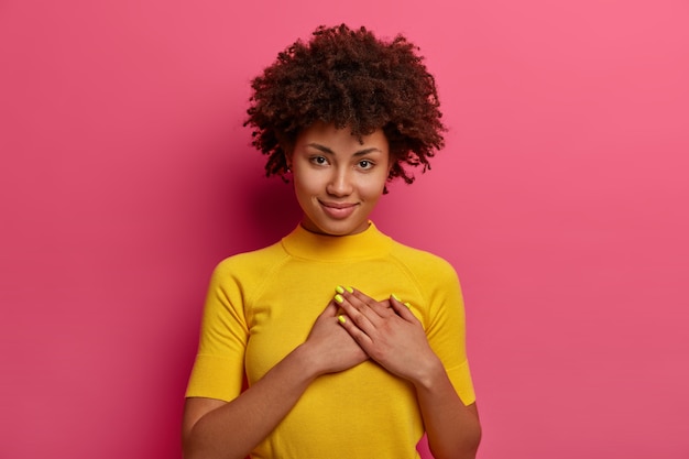 Muestra de cariño y admiración. Una mujer bonita y rizada presiona las palmas contra el corazón, agradecida por el regalo, usa una camiseta amarilla, posa sobre una pared rosada, dice que siempre estás en mi corazón, sonríe suavemente