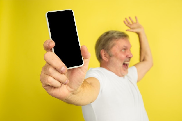 Muestra la pantalla en blanco del teléfono, apuntando hacia arriba. Retrato de hombre caucásico sobre fondo amarillo de estudio. Hermoso modelo masculino en camisa blanca.