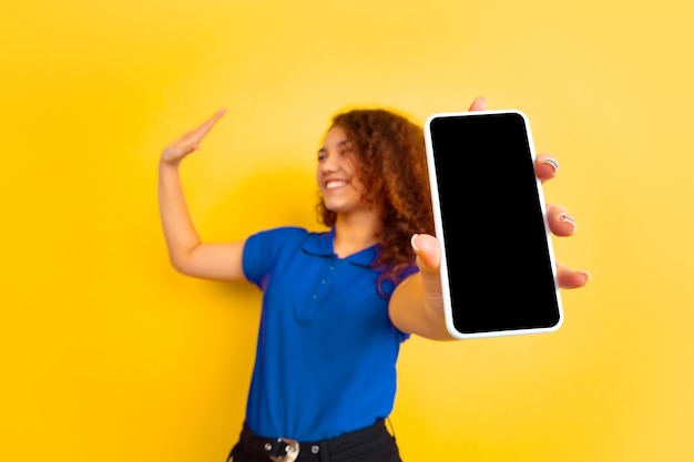 Muestra la pantalla del teléfono. Retrato de la muchacha del adolescente caucásico en la pared amarilla. Modelo rizado femenino hermoso en camisa. Concepto de emociones humanas, expresión facial, ventas, publicidad, educación. Copyspace.