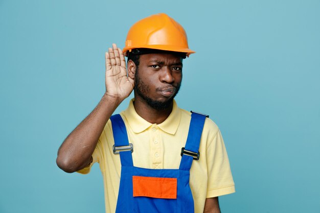 Muestra sospechosa escuchar gesto joven constructor afroamericano en uniforme aislado sobre fondo azul.