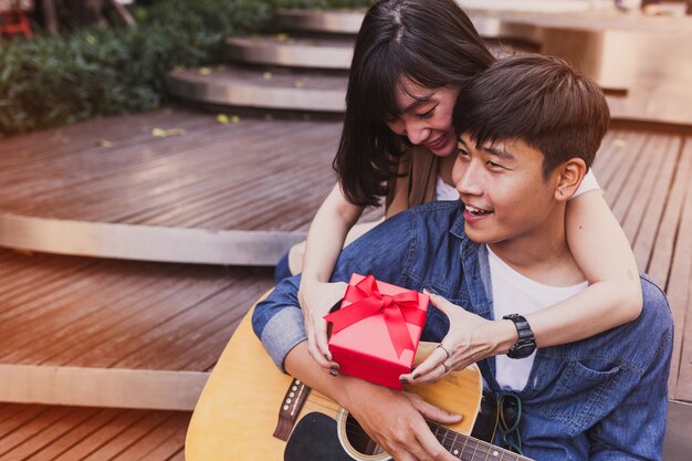 Mujer abrazando a un hombre y sosteniendo un regalo mientras éste toca la guitarra
