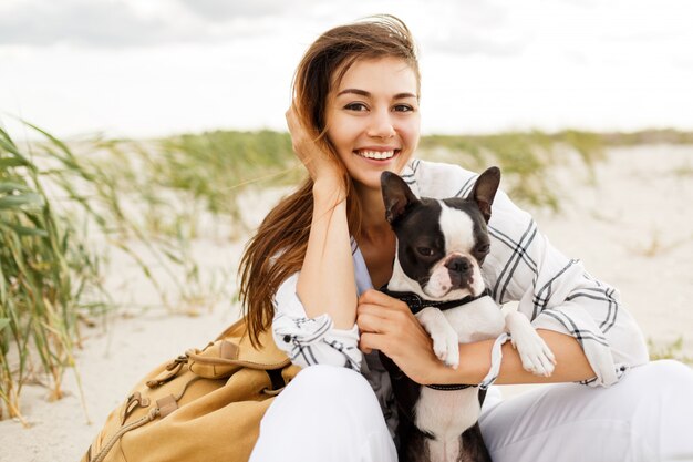 Mujer abrazando a su bulldog en la playa a la luz del atardecer, vacaciones de verano. Chica elegante con perro gracioso descansando, abrazándose y divirtiéndose, momentos lindos.