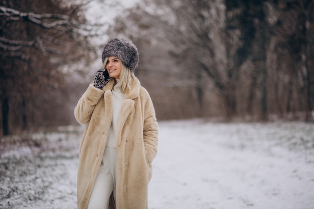 Mujer en abrigo de invierno caminando en el parque lleno de nieve hablando por teléfono
