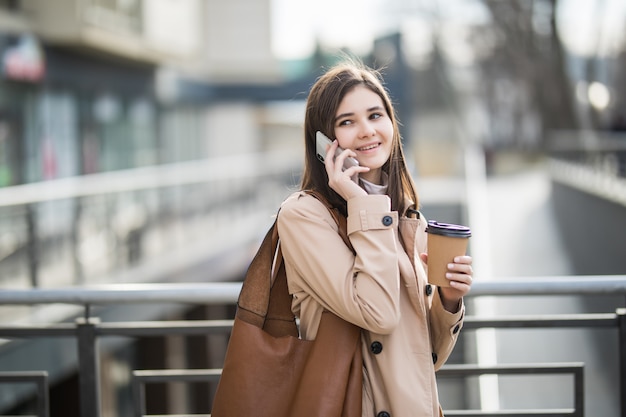 Mujer en abrigo ligero caminando por la calle con taza de café y teléfono