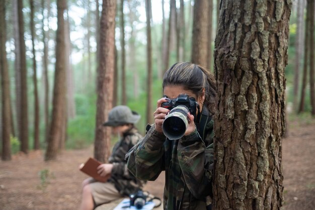 Mujer absorta con cámara en el bosque. Madre con abrigo con cola de caballo tomando fotos. hijo borroso en el fondo. Naturaleza, concepto de ocio.